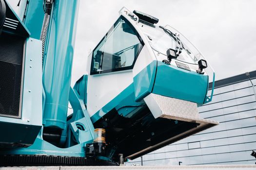 a cab with the operator of a Large blue car crane that stands ready to operate on hydraulic supports on a platform next to a large modern building. The largest truck crane for solving complex tasks