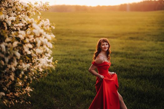 a girl in a red dress with red lips stands next to a large white flowering tree At sunset.