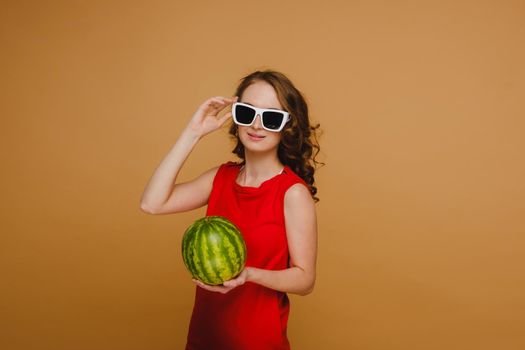 A beautiful girl in glasses and a red dress holds a watermelon in her hands.