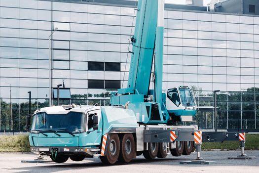 A large blue truck crane stands ready to operate on hydraulic supports on a platform next to a large modern building. The largest truck crane for solving complex tasks