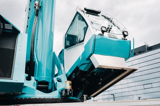 a cab with the operator of a Large blue car crane that stands ready to operate on hydraulic supports on a platform next to a large modern building. The largest truck crane for solving complex tasks