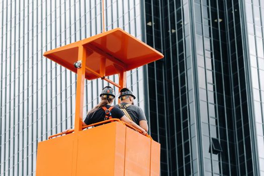 Workers in a construction cradle climb on a crane to a large glass building.The crane lifts the workers in the car seat.Construction.