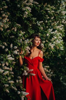 a girl in a red dress with red lips stands next to a large white flowering tree At sunset.