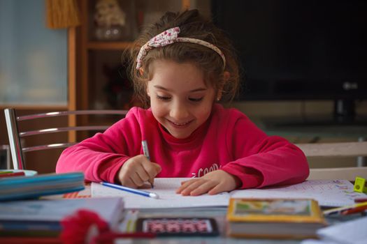 Smiling 6 year old girl does her homework, on the table the case with the colors and pens