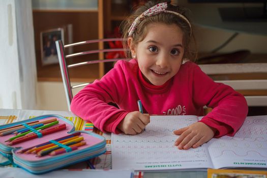 Smiling 6 year old girl does her homework, on the table the case with the colors and pens