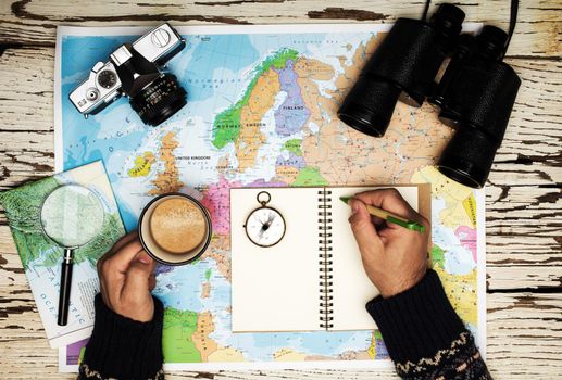 Flat lay of the travel planning concept. Top view of man hands wriìting on a diary, on the table binoculars, compass, retro photo camera, coffee, and Europe map on a white wooden table