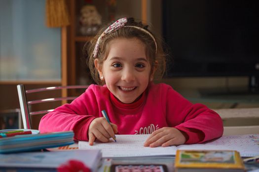 Smiling 6 year old girl does her homework, on the table the case with the colors and pens