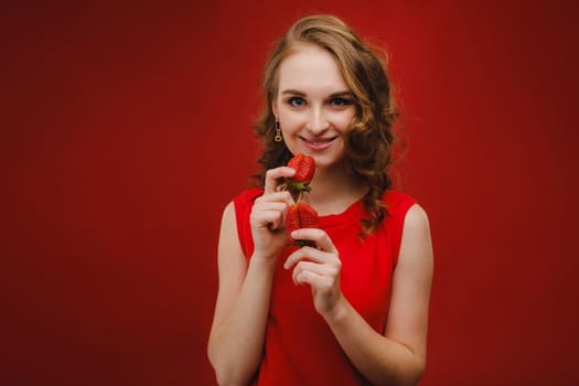 A beautiful girl in a red dress on a red background holds a strawberry in her hands and smiles.