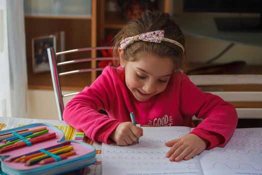 Smiling 6 year old girl does her homework, on the table the case with the colors and pens