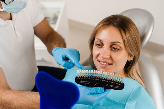 Teeth whitening. Dentist working with teeth color shades guide. Patient looking in mirror. Dentistry. Doctor checking teeth color matching samples in dental clinic