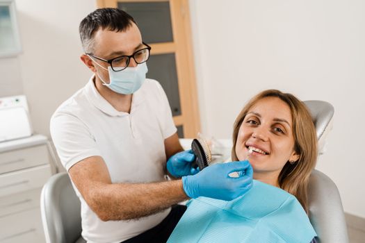 Tooth whitening. Dentist working with teeth color shades guide. Patient looking in mirror. Dentistry. Doctor checking teeth color matching samples in dental clinic