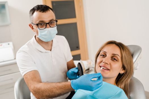 Tooth whitening. Dentist working with teeth color shades guide. Patient looking in mirror. Dentistry. Doctor checking teeth color matching samples in dental clinic