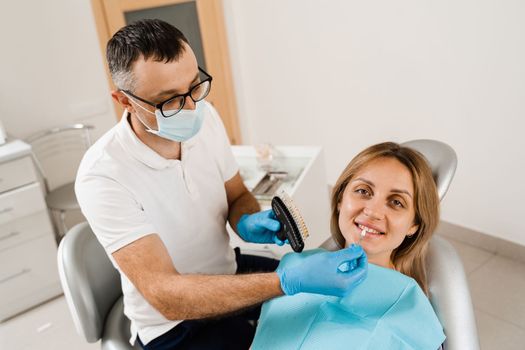 Tooth whitening. Dentist working with teeth color shades guide. Patient looking in mirror. Dentistry. Doctor checking teeth color matching samples in dental clinic