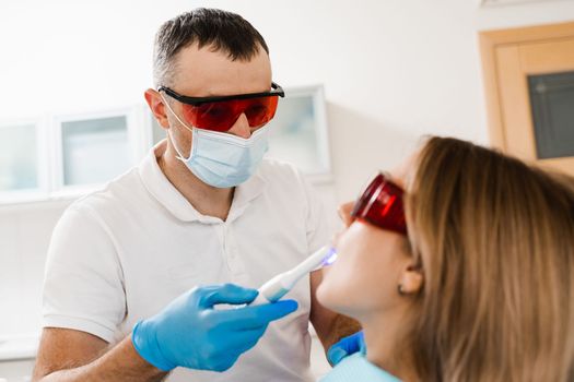 Uv illumination of photopolymer tooth filling for woman in dentistry. Dentist in red protective glasses treats and removes caries of girl patient