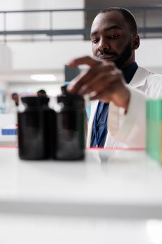 African american pharmacist taking medication bottles from drugstore shelf, selective focus on man. Pharmacy medic putting pills close up, vitamins selling, chemist front view
