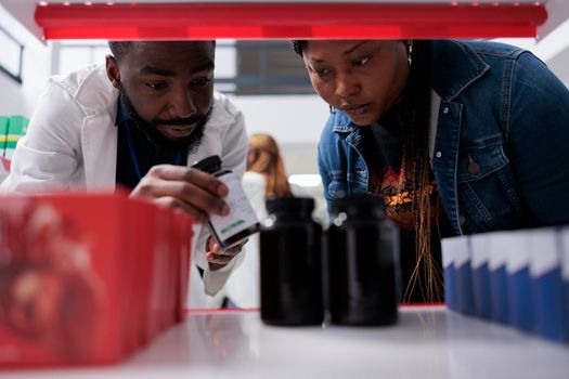 Pharmacist helping customer choosing pills on drugstore shelf, close up selective focus. African american woman taking tablets packages, pharmaceutical service, medicaments buying