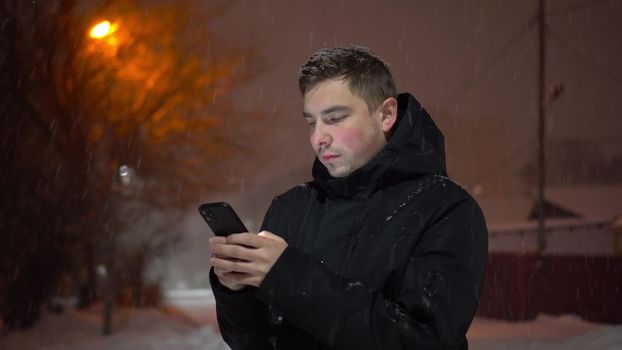 Young man on the street in winter with a phone in his hands. A man under snowfall chatting on a smartphone on a winter evening. 4k