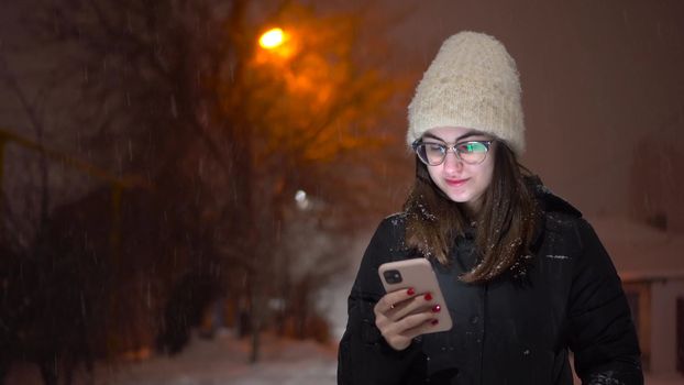 A young woman is texting on the phone on a snowy street. A girl in glasses with a phone in her hands under a snowfall. 4k