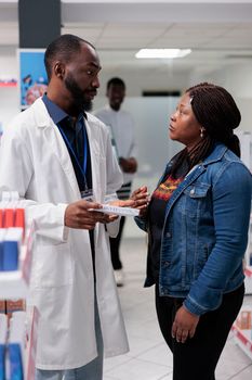 Pharmacy customer buying medication, pharmacy consultant recommending buyer pills, side view. African american woman talking with man pharmacist in drugstore aisle, all black team
