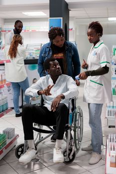 Pharmacist explaining african american customer in wheelchair vitamins instruction in drugstore. Man with physical impairment and social worker buying nutritional supplements, all black team