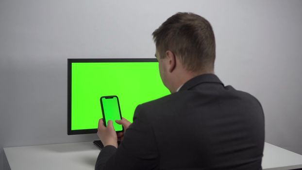 A young businessman sits at a computer and a phone in his hands with green screens. A man works in an office in a jacket. View from the back. 4k