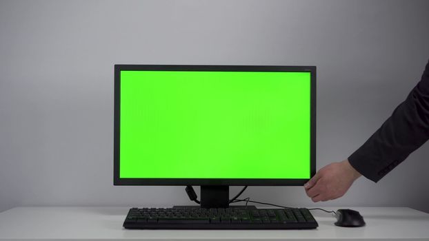 Turning on the monitor with a green screen. A man with his hand turns on the computer screen on the desktop in the office. Chroma key. 4k