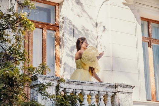 a beautiful smiling and kind woman in a gorgeous yellow dress stands on the balcony of an old vintage house.