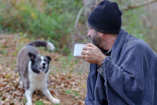 bearded man covered with a blanket drinking a cup of coffee with his dog in the background