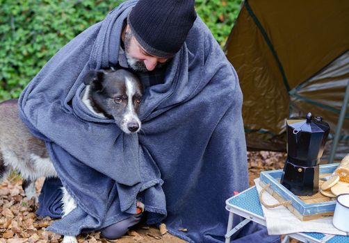 man with a beard and black camping cap covered with a blanket with his dog