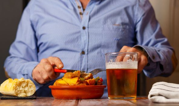 front view of man in blue shirt eating an earthenware casserole with meatballs, french fries and beer