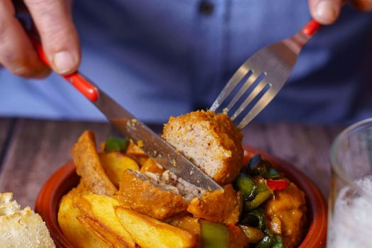 man in blue shirt cutting a meatball with fork and knife into a portion in an earthenware dish