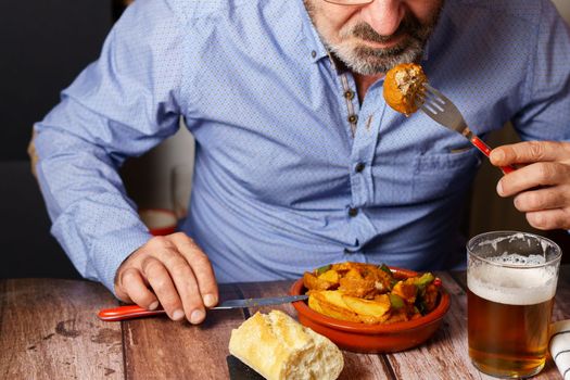 man with a beard, glasses and blue shirt eating meatball casserole with french fries and a glass of beer