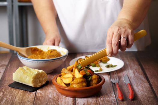 unrecognizable woman in white apron serving potatoes and vegetables over a portion of meatballs in an earthenware dish