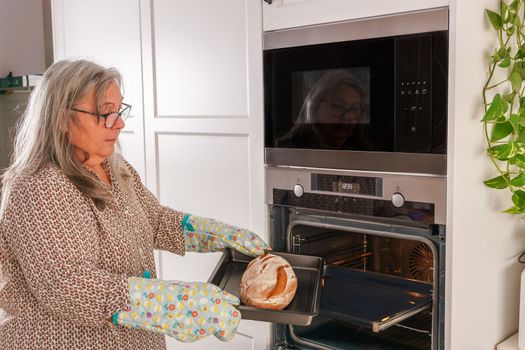 older white-haired woman putting bread in the oven in her kitchen at home