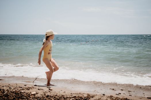 Cute little girl running along the seashore against a clear blue sea and rejoices in the rays of the summer sun. Beautiful girl in yellow swimsuit running and having fun on tropical beach
