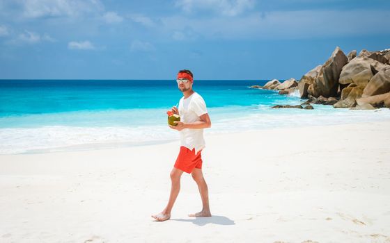 Young men in swim short with a coconut drink on a tropical beach La Digue Seychelles Islands.