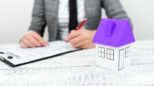 Businesswoman sitting and writing in notebook. Paper house on desk.