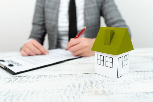 Businesswoman sitting and writing in notebook. Paper house on desk.