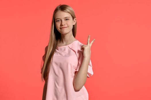 Studio portrait of a lovely blonde young lady with a long hair, in a rosy t-shirt, standing against a pink background in various poses. She expresses different emotions posing right in front of the camera, smiling and showing two fingers.