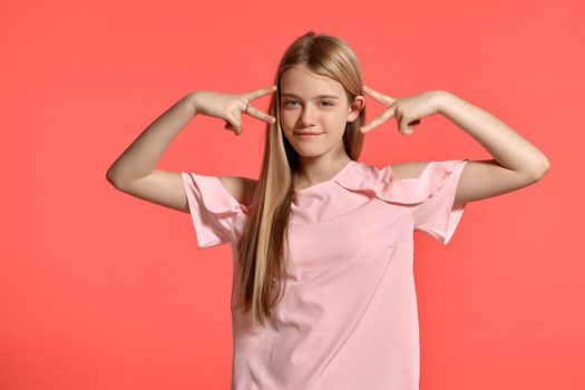 Studio portrait of a lovely blonde schoolgirl with a long hair, in a rosy t-shirt, standing against a pink background in various poses. She expresses different emotions posing right in front of the camera, smiling and looking happy.