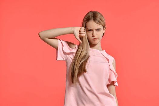 Studio portrait of a charming blonde teenager with a long hair, in a rosy t-shirt, standing against a pink background in various poses. She expresses different emotions posing right in front of the camera, looking unhappy and showing thumb down.