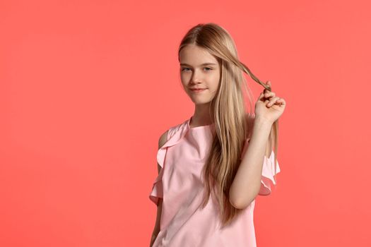 Studio portrait of a beautiful blonde teenage girl with a long hair, in a rosy t-shirt, standing against a pink background in various poses. She expresses different emotions posing right in front of the camera, smiling and winding hair on a finger.