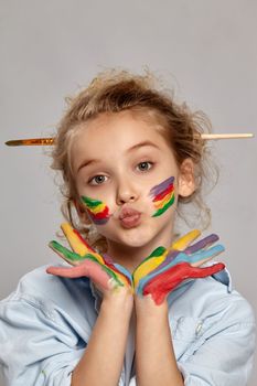 Close-up shot of a cheerful kid with a painted cheeks having a brush in her stylish hairstyle, wearing in a blue shirt and white t-shirt. She is giving a kiss at the camera, her arms are painted too, on a gray background.