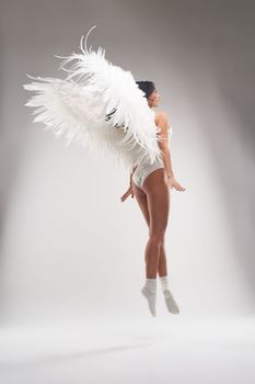 Back view of sensual woman in white socks and glittering lingerie with soft white wings floating above ground in white studio