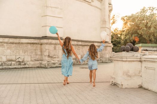 Daughter mother run holding hands. In blue dresses with flowing long hair, they hold balloons in their hands against the backdrop of a sunset and a white building