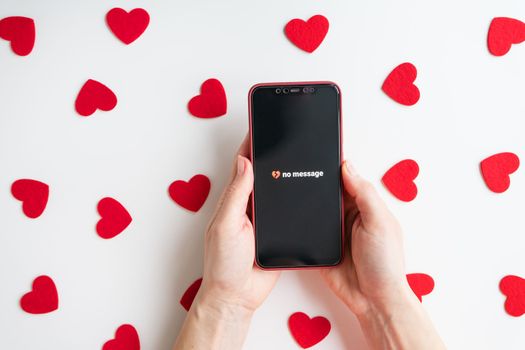 On a white table against the background of small red hearts, a girl is holding a smartphone with an activated screen and no SMS message in the form of a broken heart. The concept of the holiday Valentine's Day, February 14