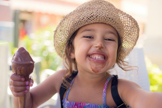 Little girl with a straw hat and a summer dress enjoys the summer heat eating a refreshing ice cream cone, it melts in her hand while she smiles funny with her mouth and nose stained with chocolate