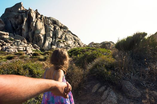 Kid strolling through the bushes holding her father's hand, in the background there are rocky mountains and the sky shines clear, subjective shot, Capo Testa, Santa Teresa di Gallura, Sardinia, Italy