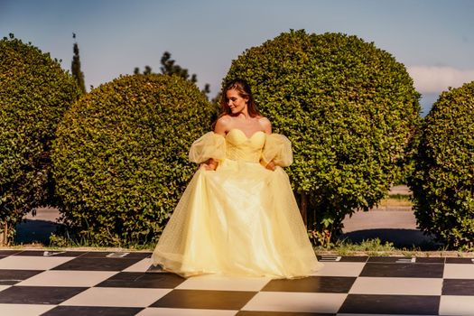 Woman yellow dress chess. A beautiful woman in a long puffy yellow dress poses on a chessboard in the park