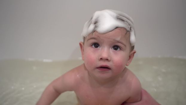 A one-year-old boy bathes in a bathtub with foam on his head. A small child washes with a small rubber duck. 4k